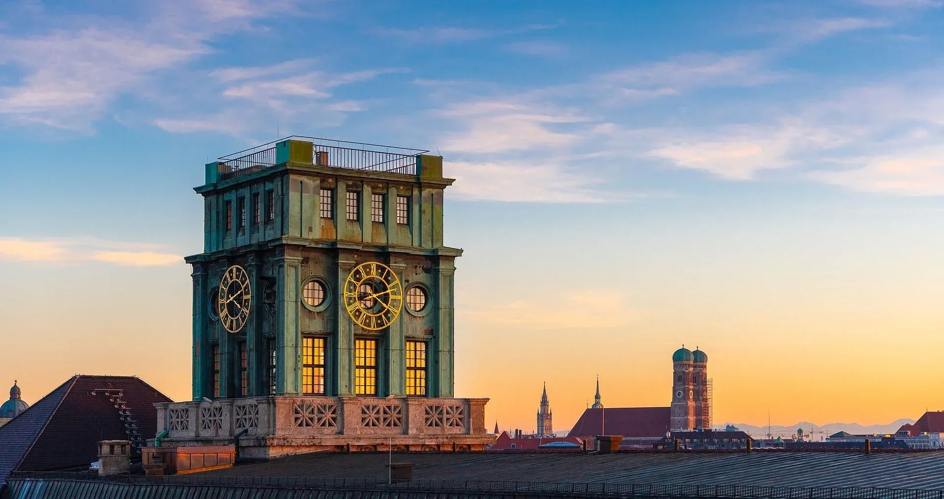 Der Thierschturm auf dem Dach des TUM-Hauptgebäudes bei Sonnenuntergang, die Zifferblätter beleuchtet, dahinter die Frauenkirche über München